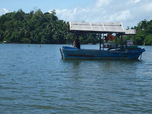 A small blue floating shop with a pitched roof sits on Madu Ganga with a wooded bank behind it.