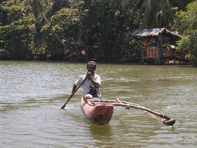 A man paddles a small outrigger canoe near a thatched waterside structure on Madu Ganga.
